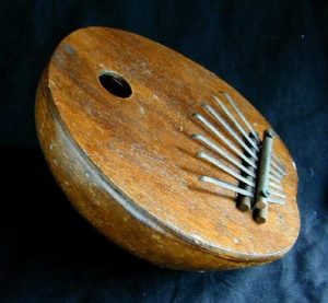 Nahaufnahme eines afrikanischen Daumenklaviers oder Kalimbas auf einem blauen Hintergrundtuch. Die Kalimba besteht aus einem rechteckigen Klangkörper mit Metallzungen unterschiedlicher Länge und Breite, die beim Anschlagen mit dem Daumen unterschiedliche Töne erzeugen.