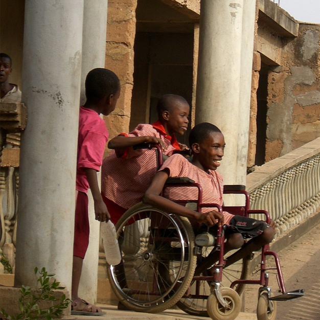 Bild von Kindern, die aus einem Gebäude kommen, während ein Kind im Rollstuhl von anderen fröhlich geschoben wird. HIFA engagiert sich für Inklusion und Rehabilitation von Kindern mit Behinderungen in Nigeria.