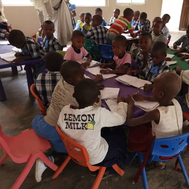 Eine Gruppe von kleinen Kindern in der Schule Uru Lokpaukwu, Afrika, sitzen auf einfachen Stühlen auf einem gebrochenen Boden. Ein kleines, süßes Mädchen steht in der Mitte und lächelt in die Kamera.