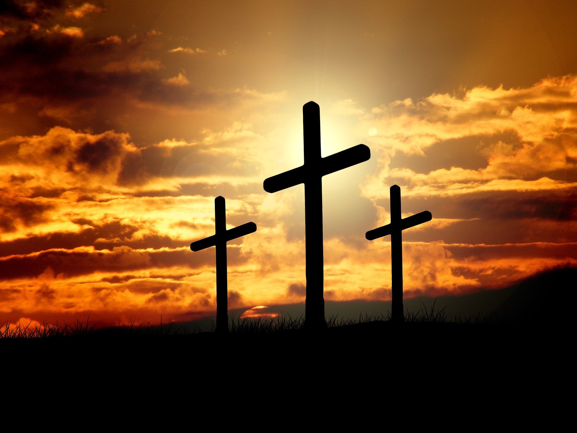 Drei Kreuze in Silhouette vor einem Sonnenuntergang, eines davon hervorgehoben. Symbolisch für den christlichen Glauben und die Opferbereitschaft von Jesus Christus.