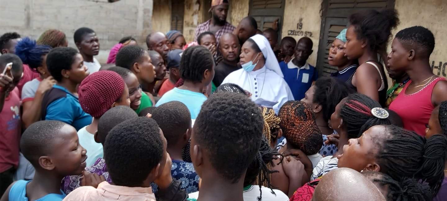 Auf dem Bild sieht man viele Menschen, die sich bei der Lebensmittelverteilung im Slumgebiet Okpoko drängeln. Dr. Joyce Ezeugo steht in der Mitte und gibt Essen an eine Frau in einem blauen Kleid. Im Hintergrund sind einige Behausungen aus Holz und Wellblech zu sehen. Die Menschen sind in einfachen, abgenutzten Kleidern gekleidet und warten geduldig auf ihre Lebensmittelration.