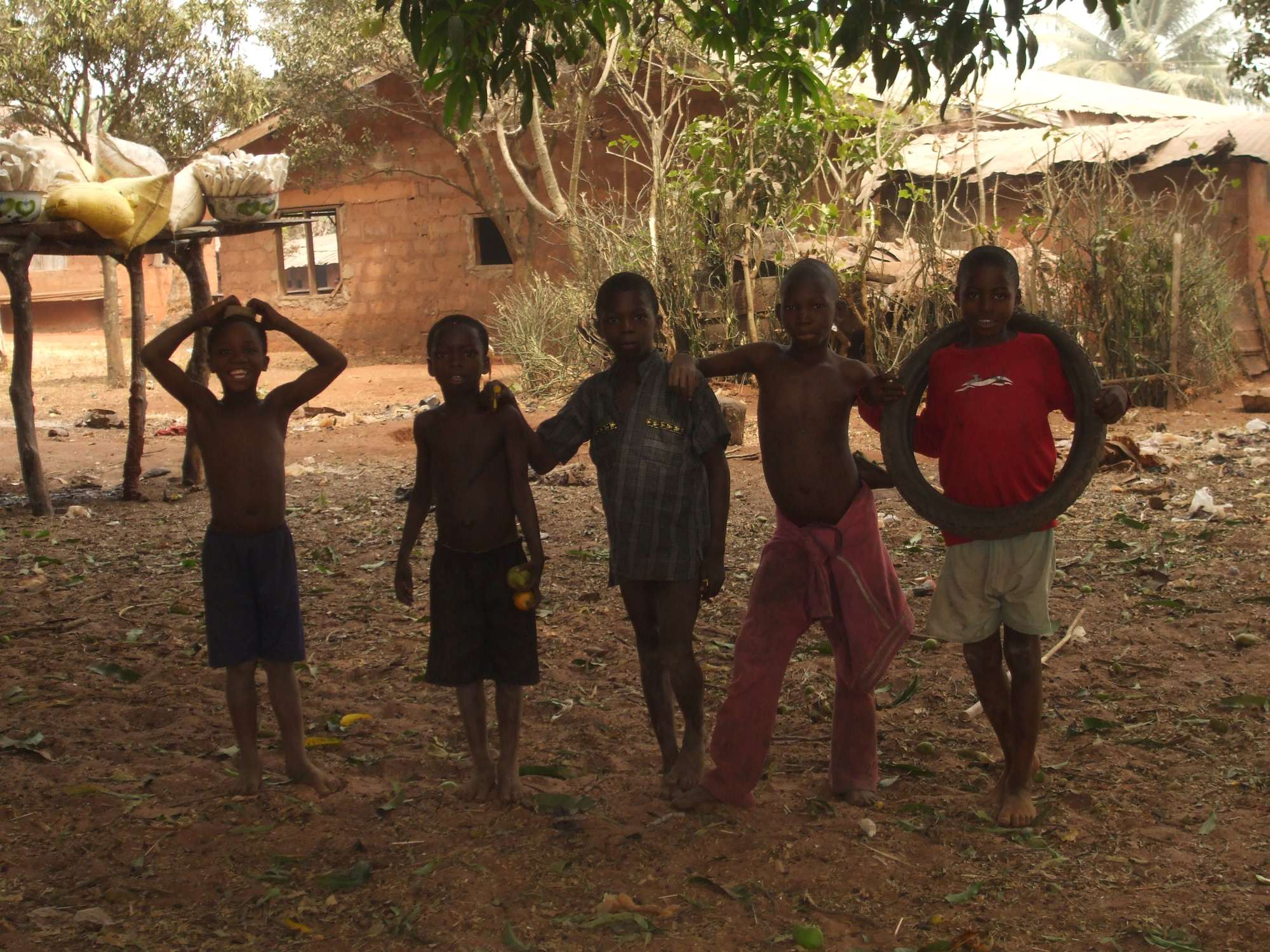 Fünf afrikanische Jungen in einem Dorf in Nigeria, lächelnd und frech, gekleidet in kurze Hosen und T-Shirts. Der Boden ist trocken und karg.