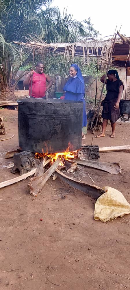 Dr. Joyce Ezeugo und ein Mann kochen das Fruchtfleisch der Palmfrüchte in einem riesigen Kessel auf einer Feuerstelle im Freien. Eine Frau kommt gerade aus dem Hintergrund an. Im Hintergrund ist eine Überdachung mit Palmblättern und eine Palme zu sehen.