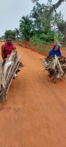 Sr. Joyce auf einer sandigen Straße, die Schubkarre mit Holz beladen, das für das Kochen von Palmkernen benötigt wird