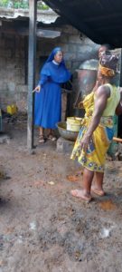 Dr. Joyce Ezeugo leert gekochte Palmfrüchte in eine alte Presse, während Palmöl herausläuft. Im Hintergrund ist ein altes Gebäude zu sehen.