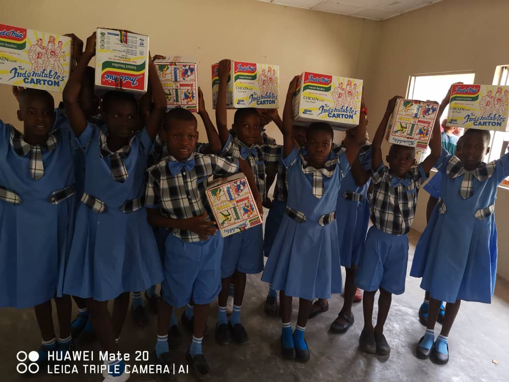Freudestrahlende Schulkinder in blauer Schuluniform halten die gespendeten Lebensmittelschachteln hoch. Symbol für Solidarität und Gemeinschaftshilfe während des Corona-Lockdowns 2022 an der Adolf Paster Present School in Uru Lokpaukwu, Abia State, Nigeria.