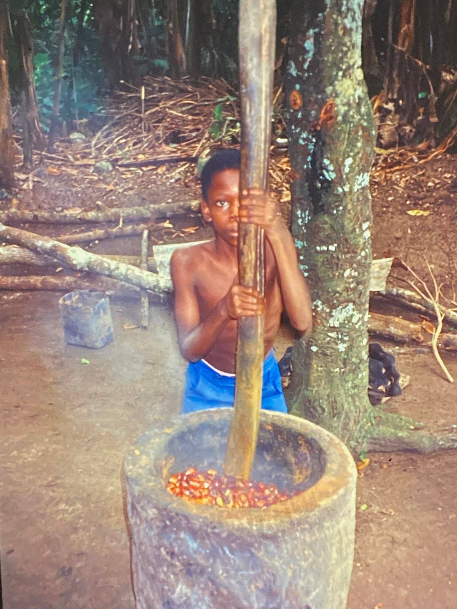 Afrikanischer Junge in kurzer blauer Hose, der vor einem großen Betonmörser steht und mit einem Holzprügel gekochtes Palmfruchtfleisch zu Brei verarbeitet.
