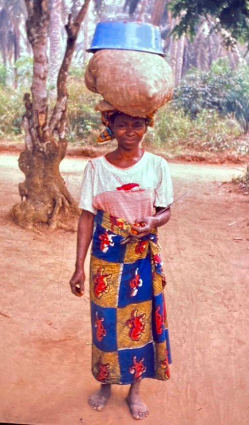 Portrait einer lächelnden Frau in afrikanischer Kleidung, die einen Korb mit Palmfrüchten auf ihrem Kopf trägt, auf dem Weg ins Dorf zur weiteren Verarbeitung.
