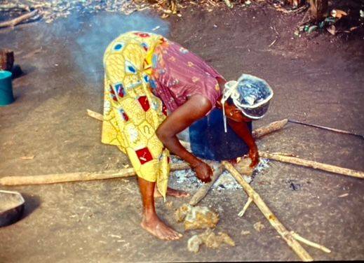 Großer Topf mit kochenden Palmfrüchten auf einer Scheiter-Kochstelle im Freien. Eine Frau legt zusätzliche Scheite auf das Feuer, um sicherzustellen, dass die Flamme nicht erlischt.