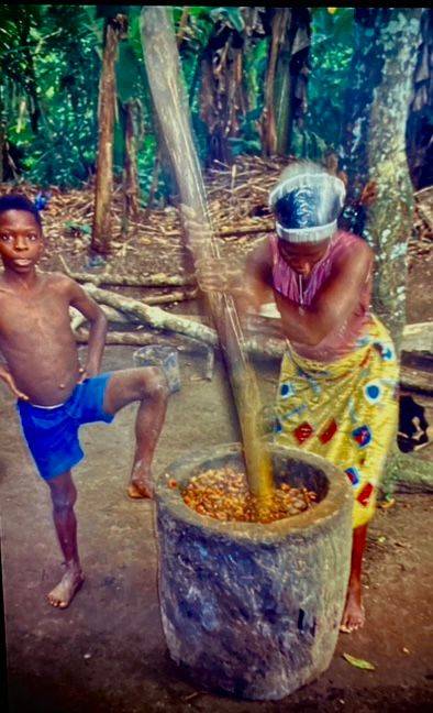 Afrikanische Frau stampft gekochtes Palmfruchtfleisch in einem großen Betonmörser zu Brei, während ein Junge in einer kurzen blauen Hose daneben steht und zuschaut.