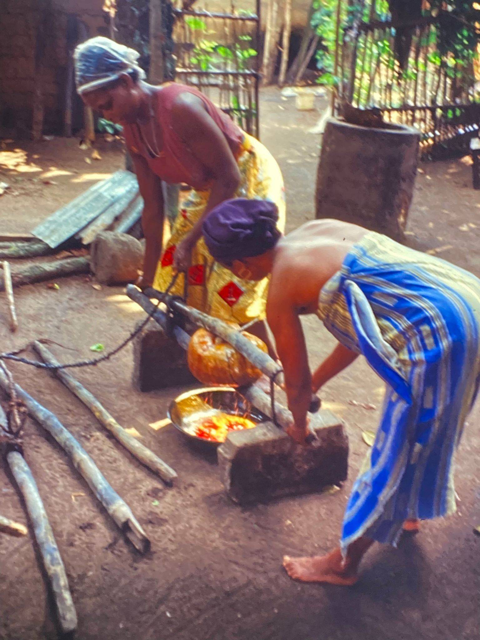 Zwei Frauen pressen gekochte Palmfasern in einer selbstgemachten Maschine, das freigesetzte Palmöl sammelt sich in einer darunter stehenden Schüssel.